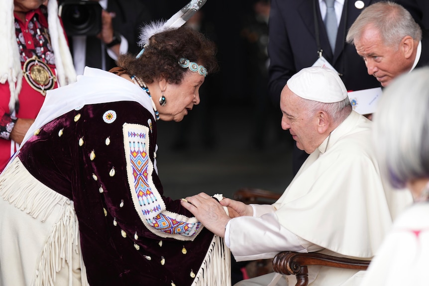 Gekleed in een gewaad en in een rolstoel schudt paus Franciscus de hand van een vrouw in traditionele klederdracht. 