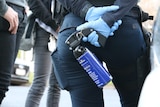A close up shot of a police officer wearing blue plastic gloves holding a blue metal bottle of capsicum spray behind her back.