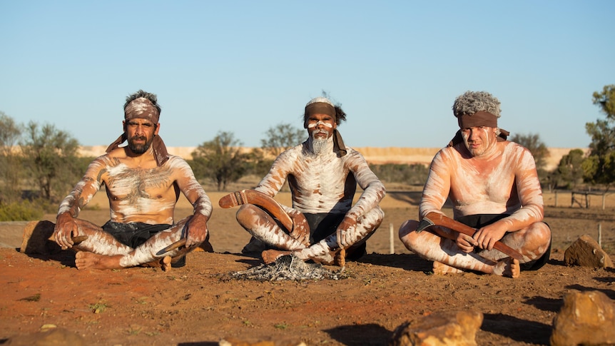 three men with ingidenous body paint look at the camera while sitting cross legged with a mine in the background
