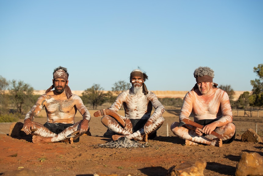 three men with ingidenous body paint look at the camera while sitting cross legged with a mine in the background