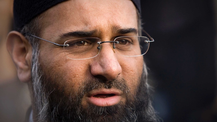 Close view of Anjem Choudary's face. He is wearing glasses and a black cap, and has a full, greying beard. He is mid-speech.