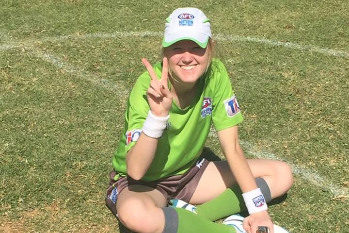 Jessie, a woman, sits on the ground crossed legged, doing a peace sign with her hands and smiling. 