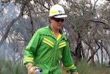 A man in a green fire suit and white helmet sets dry grass alight in the bush.