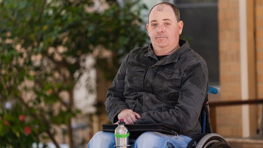 A man in a black shirt and jeans in a wheelchair in front of a building with a tree in the background.