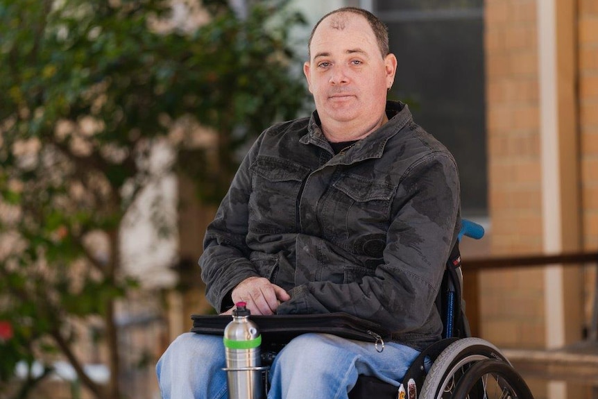 A man in a black shirt and jeans in a wheelchair in front of a building with a tree in the background.