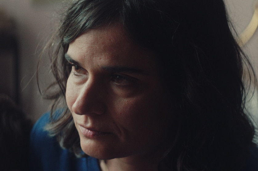 A middle aged woman with shoulder length dark hair and neutral expression sits with child on lap lit by natural light.