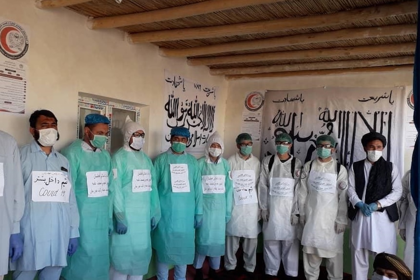 Men in surgical gowns and medical masks stand before posters in Afghan language promoting a coronavirus safety campaign