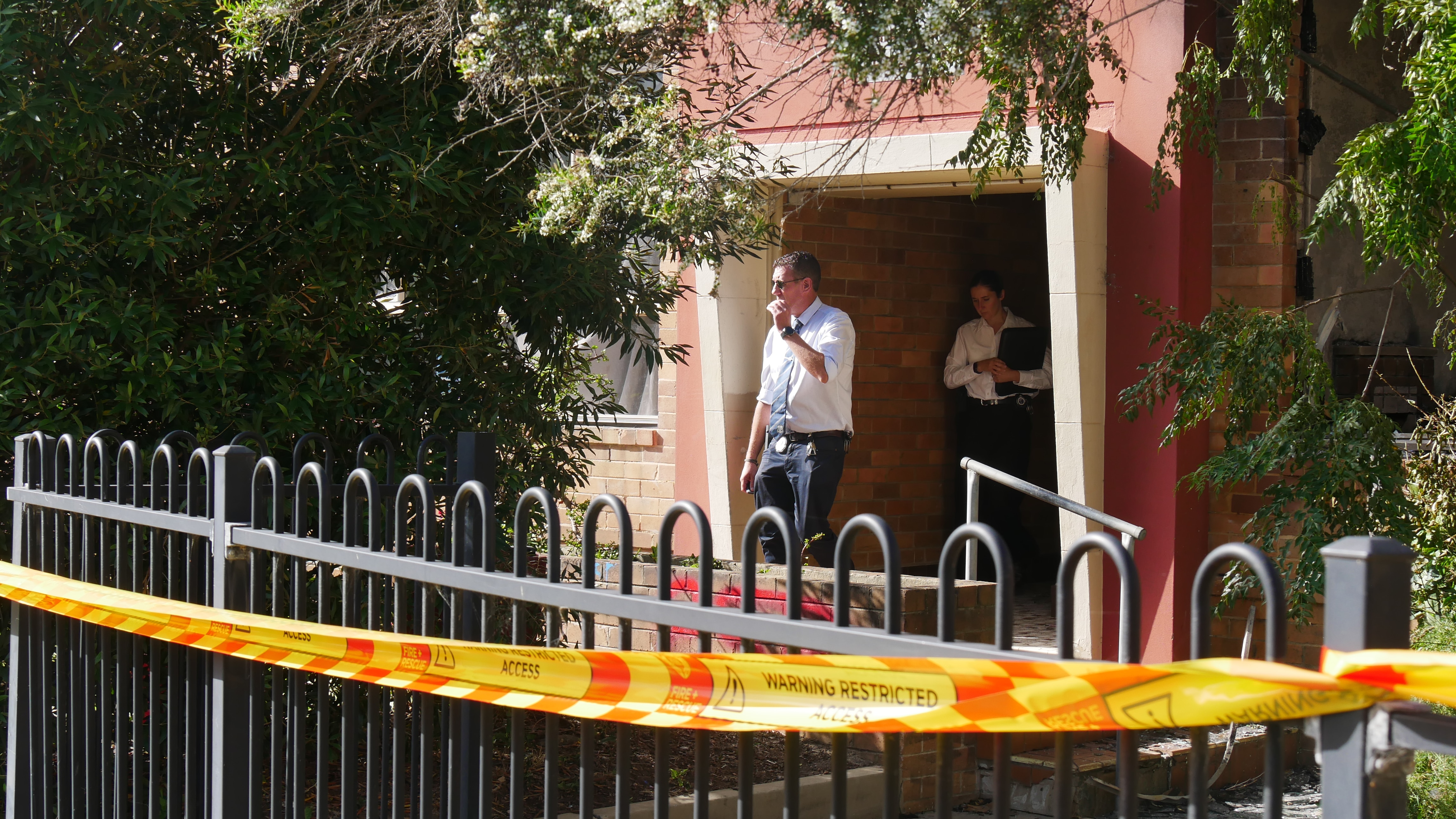 Um homem de camisa branca, gravata azul, uma mulher de camisa branca, segurando uma pasta, sai por um portão com fita policial por cima da cerca.