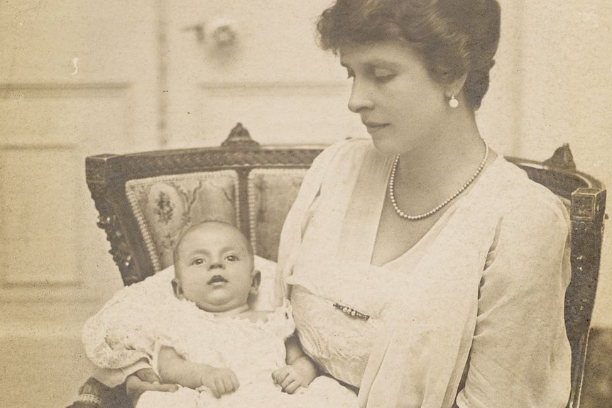 A sepia photograph of baby Philip wearing a white dress, in his mothers arms as she sits in a chair.