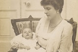 A sepia photograph of baby Philip wearing a white dress, in his mothers arms as she sits in a chair.