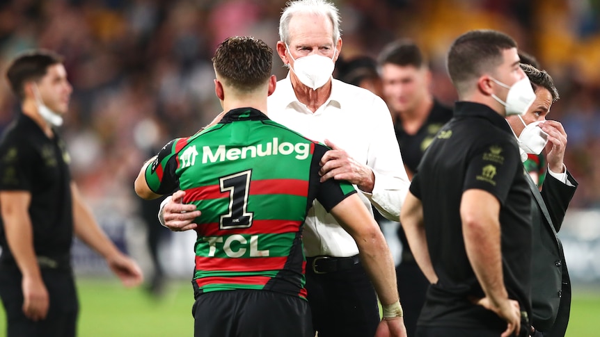 Wearing a face mask, Wayne Bennett hugs Souths' Blake Taaffe on the field.