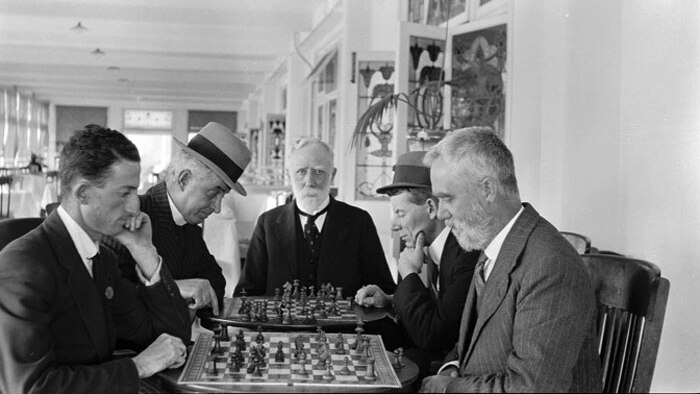 Five men in suits around a table playing chess in a cafe