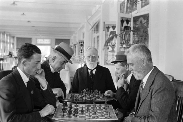 Five men in suits around a table playing chess in a cafe