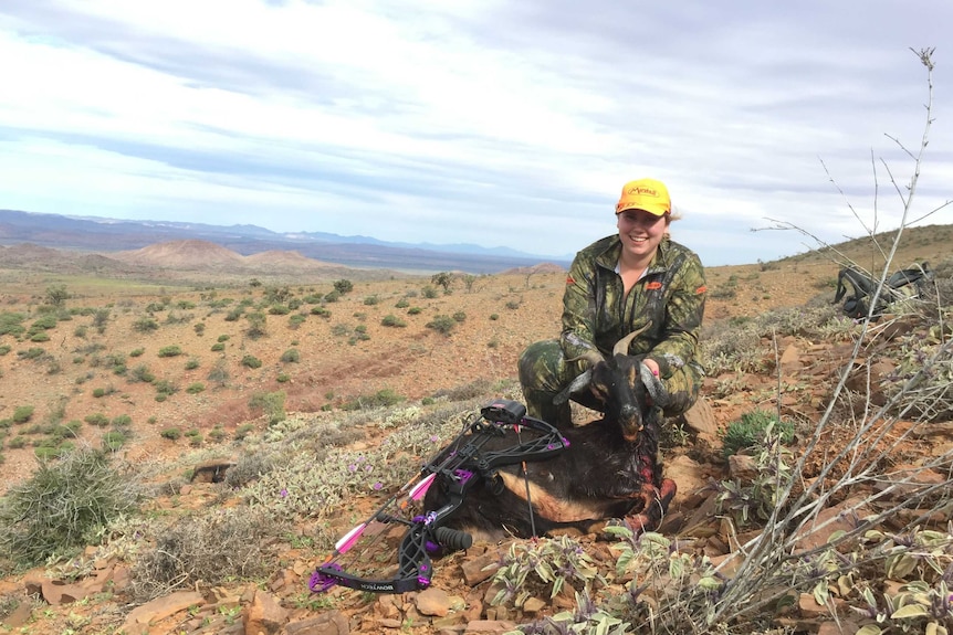 A woman in camouflage kneeling behind a dead goat.