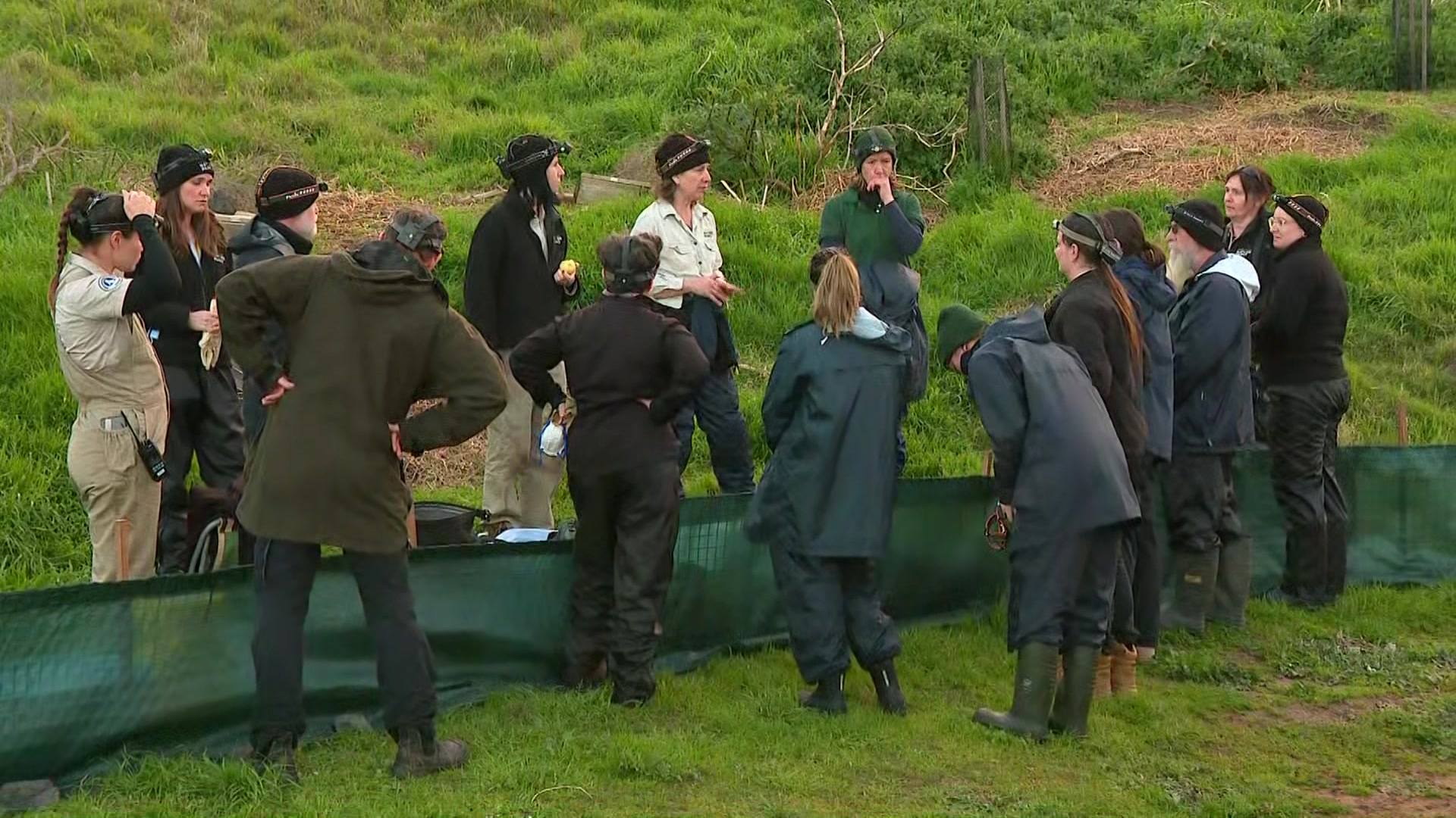 People dressed in coats and wearing beanies gather on either side of a small green material divider stretched across grass.