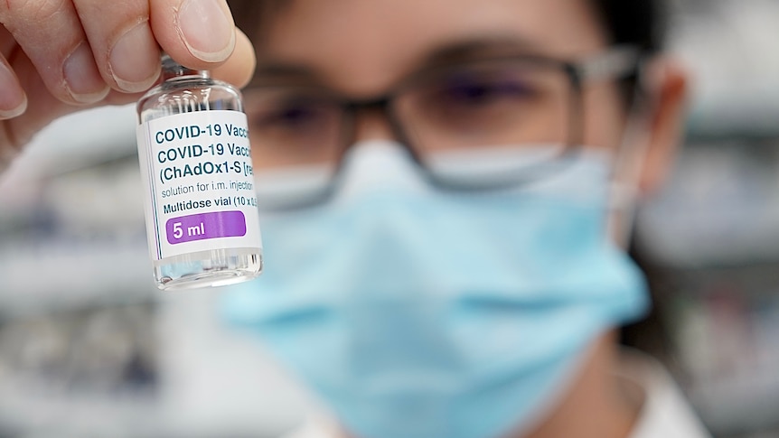 A woman in a mask with glasses holds up a vial of vaccine.