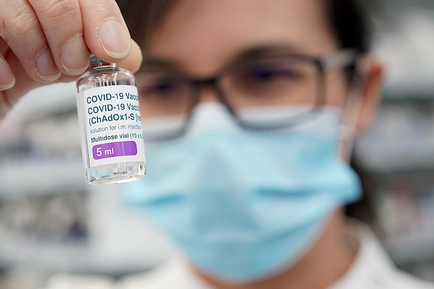 A woman in a mask with glasses holds up a vial of vaccine.