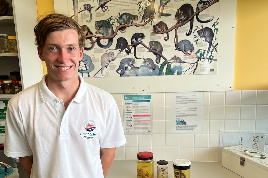 A teenage boy stands in a science classroom wearing a white shirt, smiling.