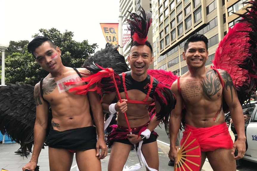 Three men wearing feathered wings and holding props celebrating Mardi Gras in Sydney.