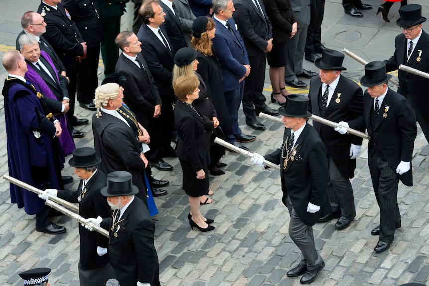 Men in top hats and tailcoats walk past Nicola Sturgeon and other ministers standing outside on cobbled stones