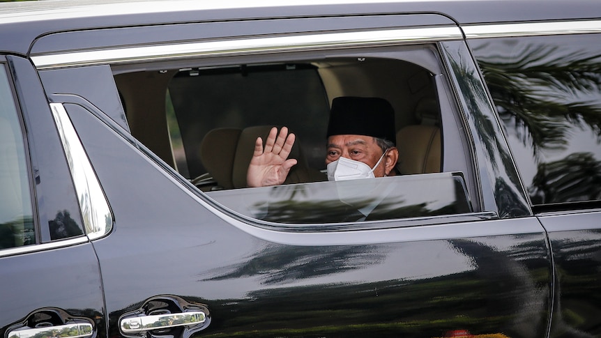 A man wearing a white disposable face mask waves from the window of a black car.