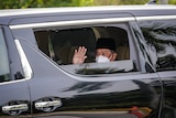 A man wearing a white disposable face mask waves from the window of a black car.