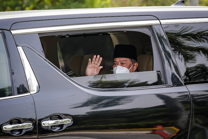 A man wearing a white disposable face mask waves from the window of a black car.