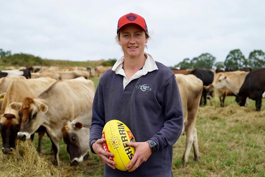 AFLW player Shelley Scott stands holding a Sherrin in front of dairy cows.