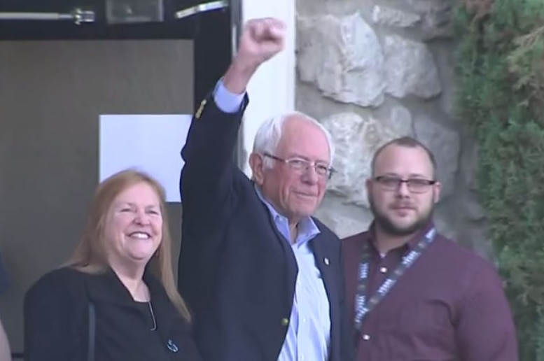 Bernie Sanders stands at the exit of a building with his fist in the air, standing by his wife and three hospital workers.