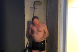 A middle-aged man in board shorts standing under a running shower connected to a garden hose