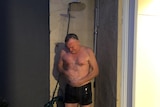 A middle-aged man in board shorts standing under a running shower connected to a garden hose