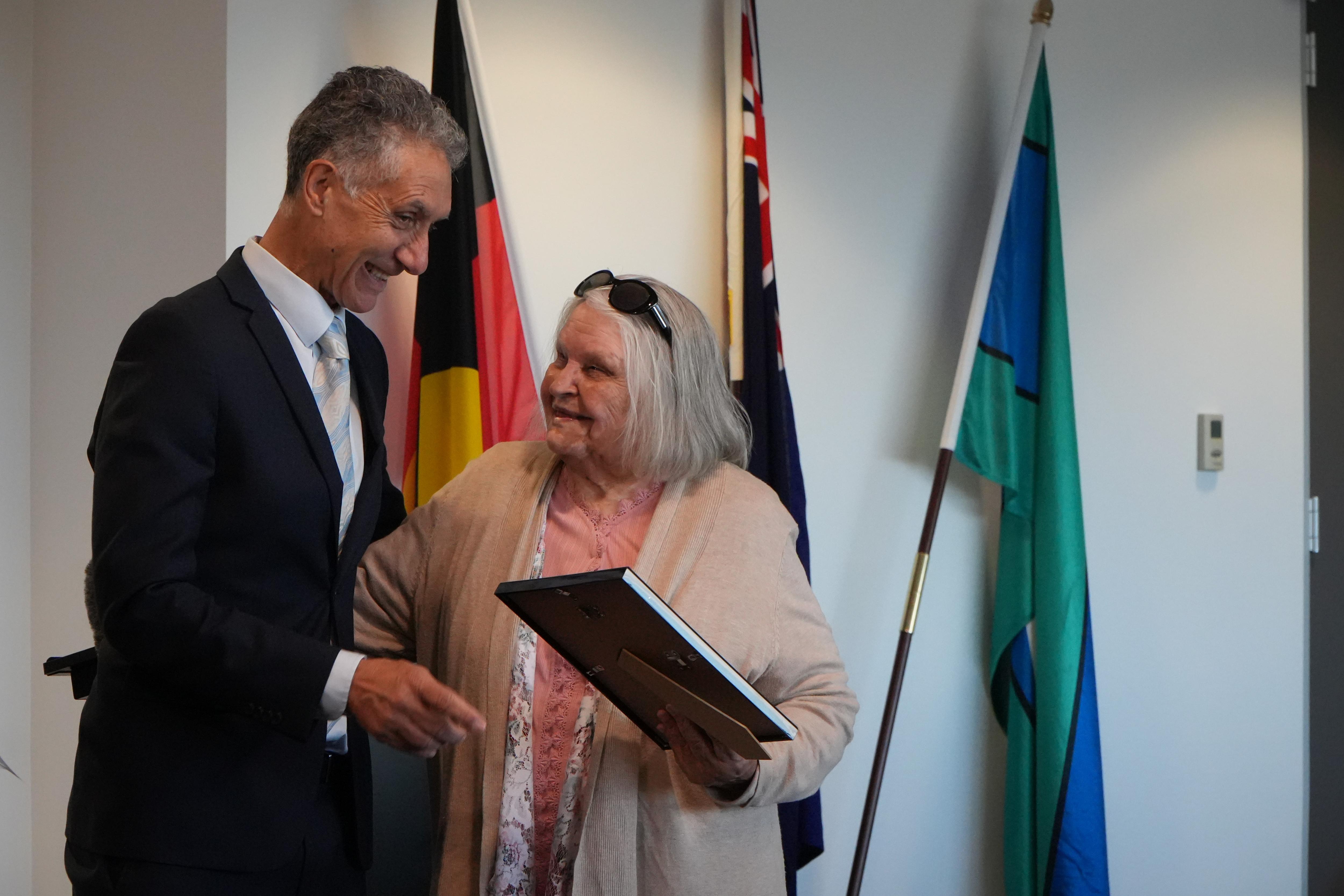 A shorter 88 year old woman smiling at a taller man in a suit as they go to embrace. She is holding a frame in her hand