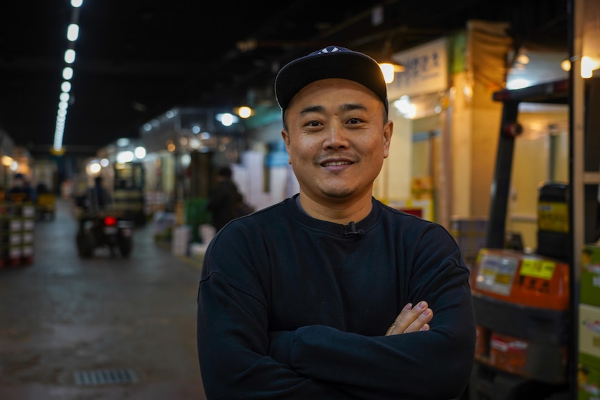 An Asian man wearing a cap and black jumper stands in front of market stalls.