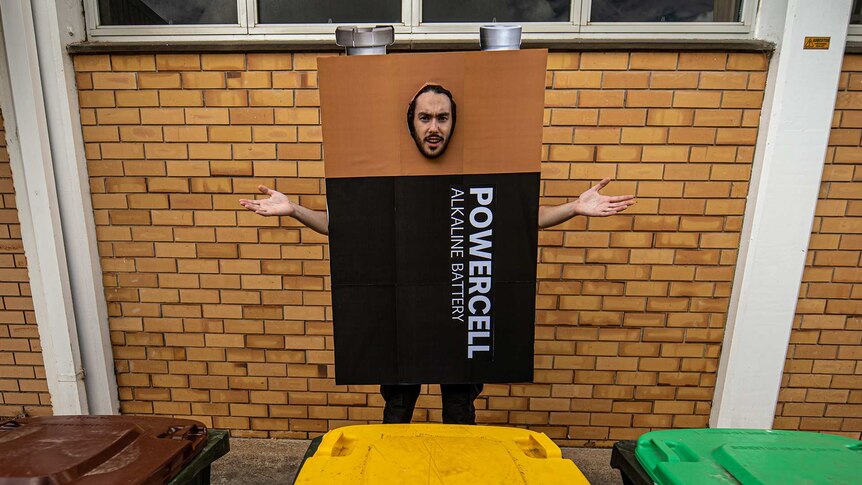Alex dressed as a battery standing confused in front of three different bins.