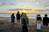 A smiling woman with a denim jacket, scarf faces the camera, while a group of men and woman watch the sunrise at the beach.