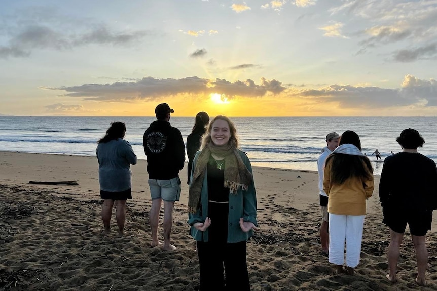 A smiling woman with a denim jacket, scarf faces the camera, while a group of men and woman watch the sunrise at the beach.