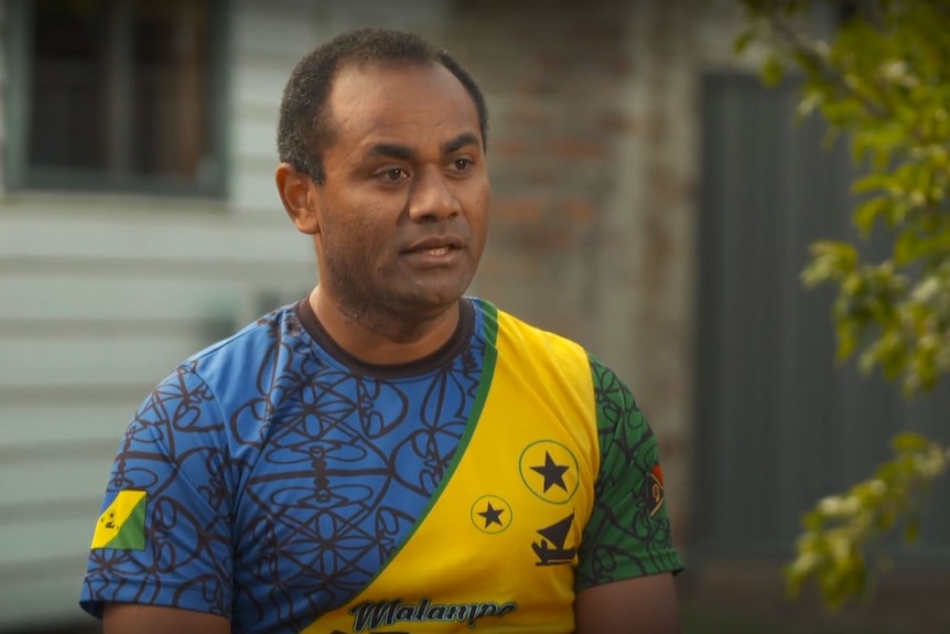 Man wearing bright coloured top speaks to camera with green tree and house behind him. 