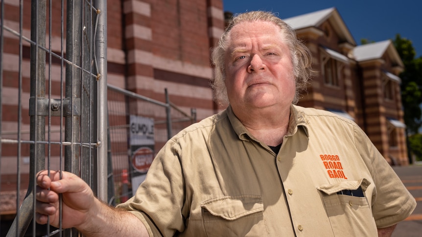 A cross man clutches an iron fence next to a building