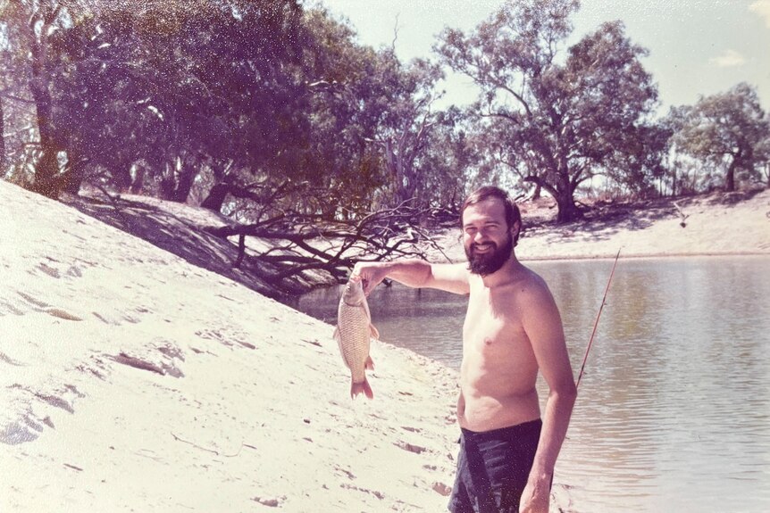 A man stands with no shirt holding a fish.