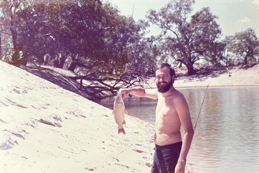 A man stands with no shirt holding a fish.