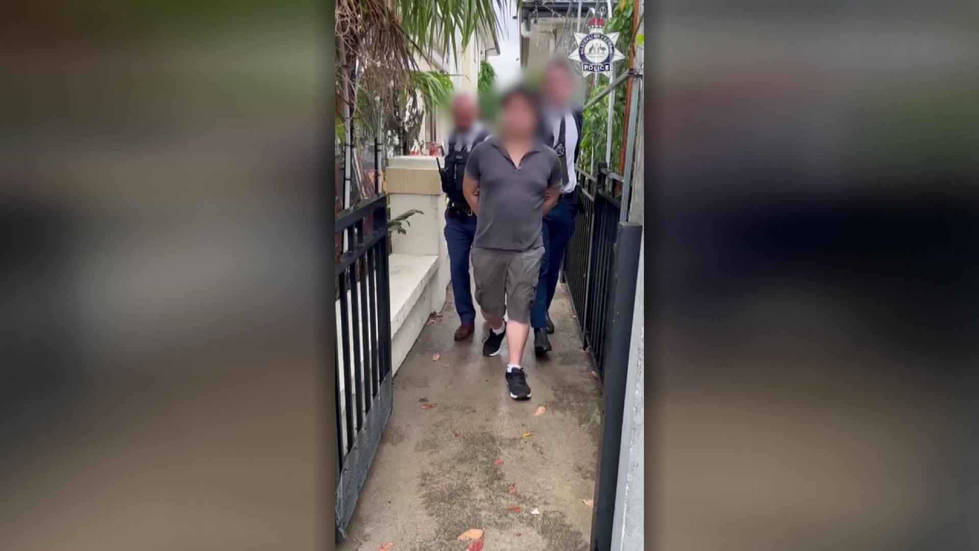 A man with his face blurred and hands cuffed behind his back is led by two police officers.