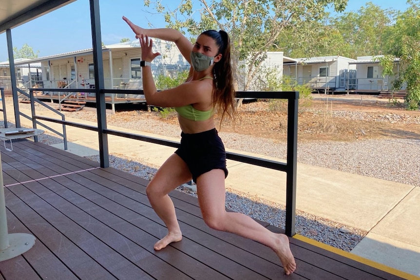 A woman wearing a mask, practising contemporary dance on a donga verandah.