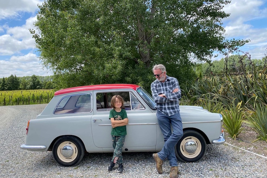 A man wearing sunglasses stands with his arms crossed, while leaning on a car, next to his grandson