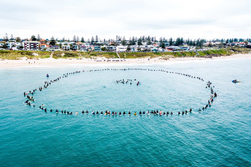 An aerial view of surfers forming a large ring around a smaller ring of surfers in the water at North Cottesloe Beach.