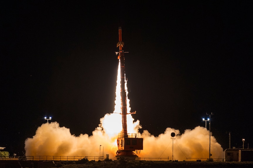 A red missile launches upwards with its orange trail visible at night.