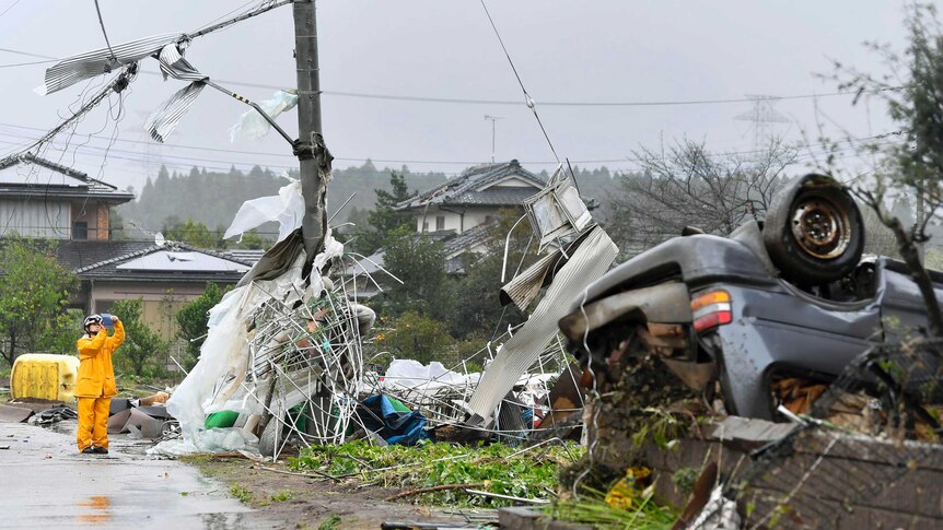 A car is upturned on the side of the road, electricity lines and poles are battered, debris lies everywhere.