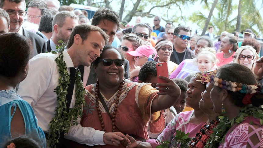 French President Emmanuel Macron wearing flowers around neck poses for selfie with Polynesian woman in sunglasses in crowd