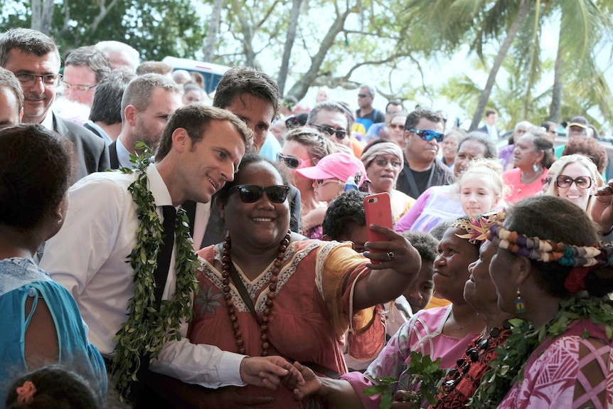 French President Emmanuel Macron wearing flowers around neck poses for selfie with Polynesian woman in sunglasses in crowd