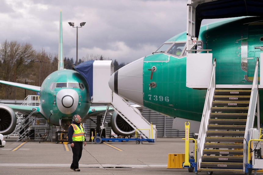 Looking from the groudn, unpainted green Boeing 737 MAX jets are parked next to each other with a small man between them.