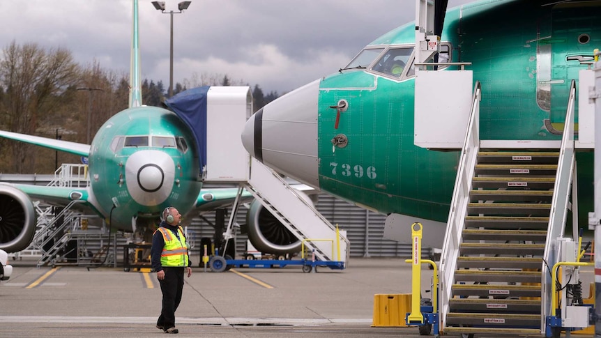 Looking from the groudn, unpainted green Boeing 737 MAX jets are parked next to each other with a small man between them.